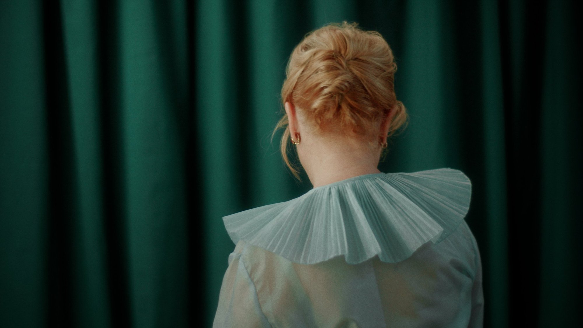 Woman from behind in a pleated pale-blue ruff against a green curtain