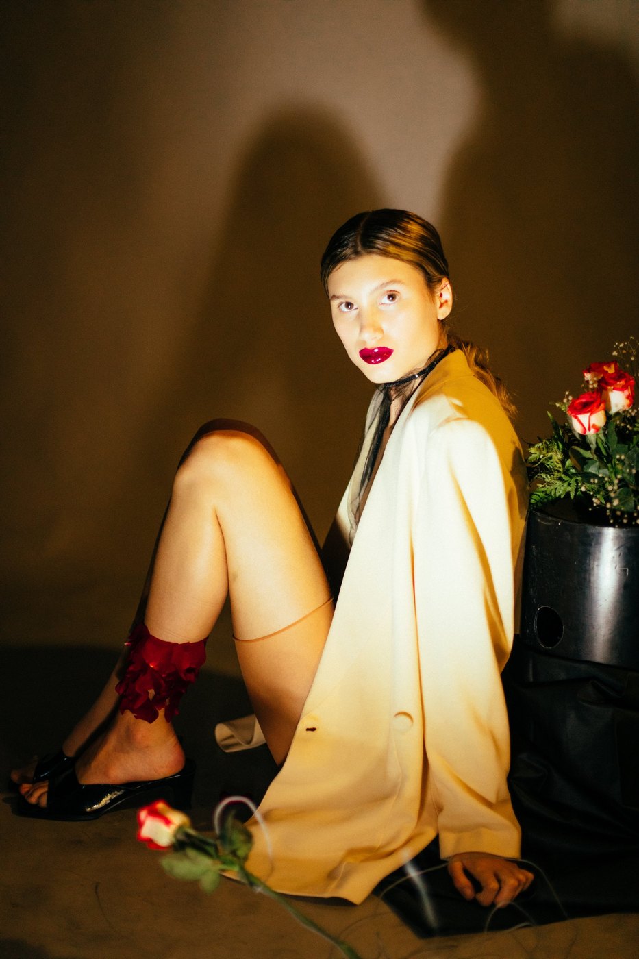 Model seated in a cream blazer with red petals on her ankle